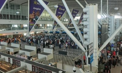 airport departure board showing cancelled flights, stranded traveller with suitcase in terminal, Qatar Airways cancellation message screen, Australian airport waiting area travellers, international flight disruption news, Joburg ETC